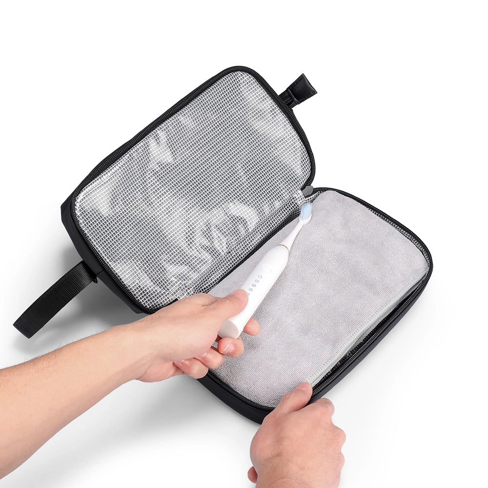 Black Switchstorage V toiletry bag with wet mesh and dry fabric compartments, showing a hand placing a white electric toothbrush in the dry section.
