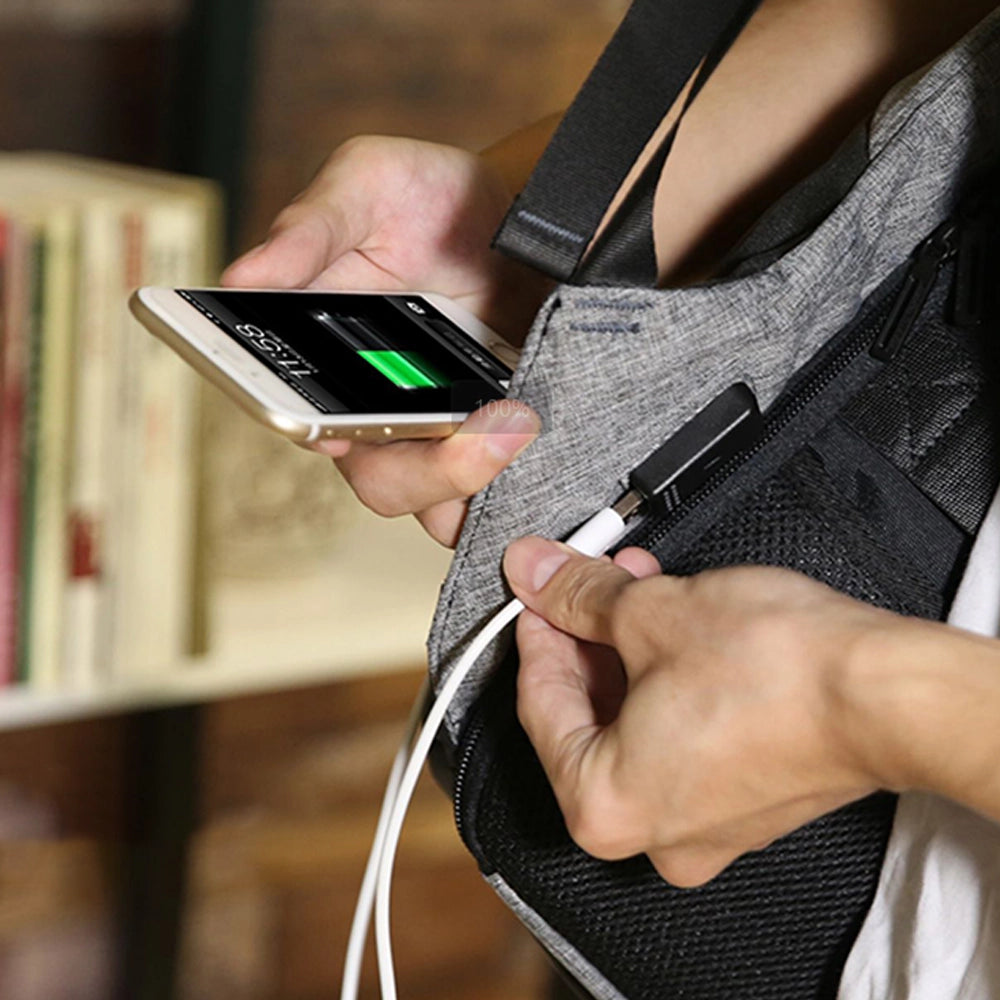 Person plugging white USB cable into gray and black Mocchasio anti-theft backpack to charge smartphone, highlighting convenient USB charging feature.