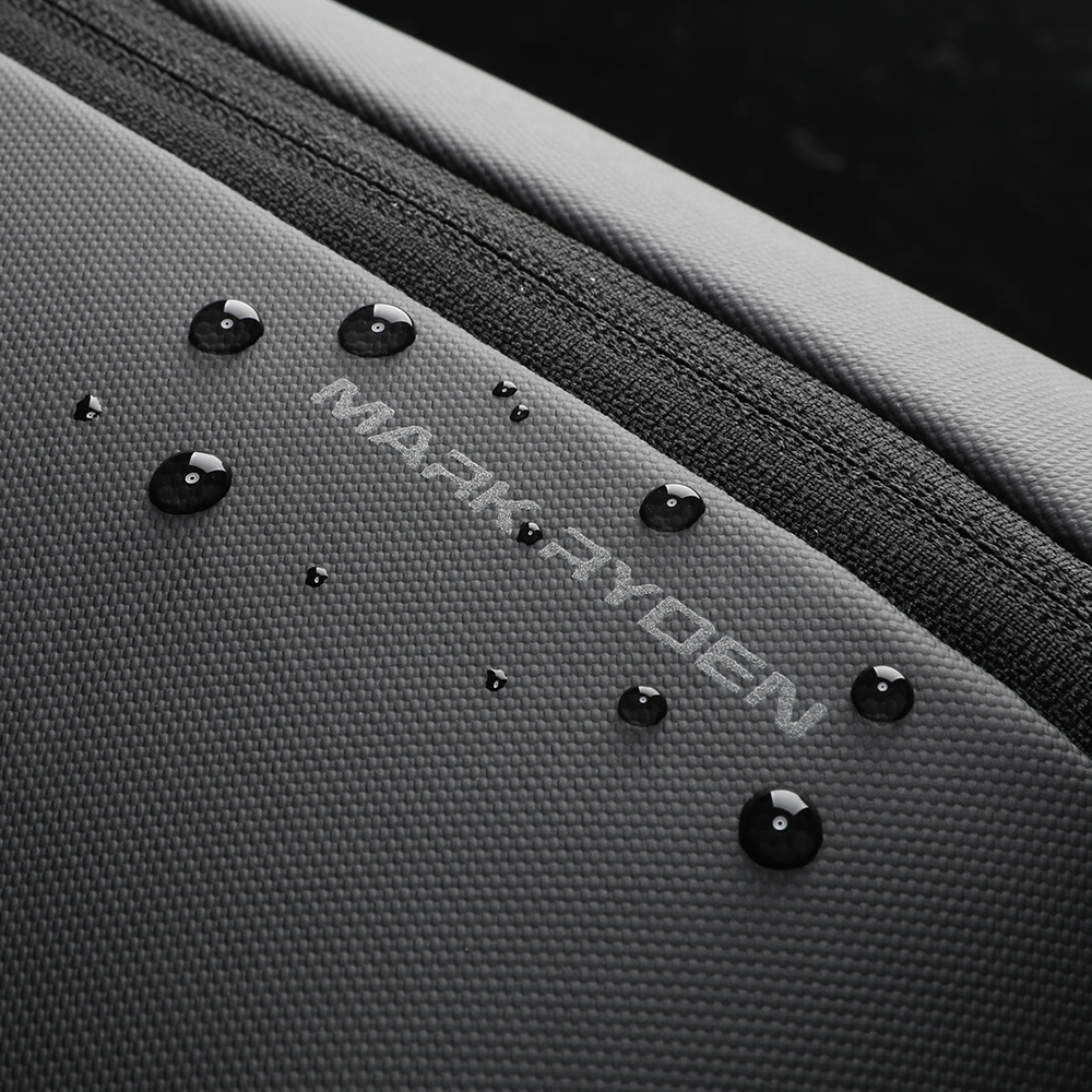 Close-up of MarkRyden Colonne high-capacity urban commuting crossbody bag with water-resistant textured fabric, embossed brand logo, and water droplets beading on surface.