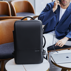 Black MarkRyden 40L three-layer main pocket backpack for business trips, travel, and college, displayed on a round table with a laptop and a person in a blue blazer nearby.